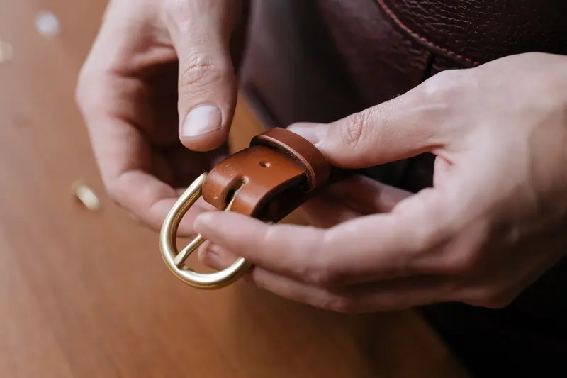 Close-up shot of hands holding a brown leather belt with a brass buckle, showcasing craftsmanship