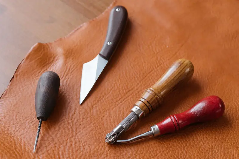Close-up of leather crafting tools including awl, knife, and groover on a brown leather sheet