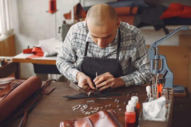 Focused male master in apron and check shirt attaching small metal snaps with tools on piece of leather while sitting at table and creating accessory in contemp
