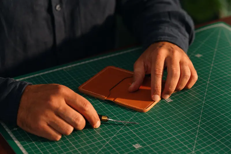 Close-up of hands crafting a leather wallet on a green cutting mat, emphasizing artisanal craftsmanship